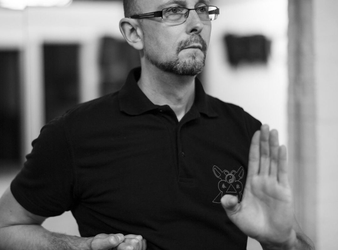 Black and white photo of Sifu Alan in a black club polo shirt, one hand raised in an open guarding palm and the other closed in a fist, eyes calm and focused.