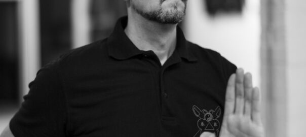 Black and white photo of Sifu Alan in a black club polo shirt, one hand raised in an open guarding palm and the other closed in a fist, eyes calm and focused.